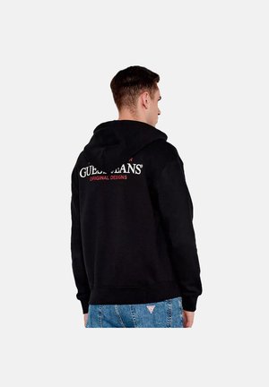 Man draagt een zwarte hoodie met de tekst "Guess Jeans Original Designs" op de rug en een blauwe spijkerbroek tegen een effen achtergrond.