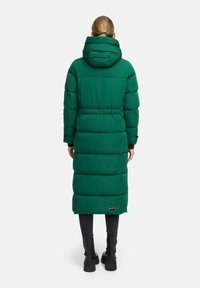 Lungo cappotto verde puffer con cappuccio, caratterizzato da sezioni trapuntate, vita cintrata e un patch con logo vicino all'orlo. Leggings neri e stivali.