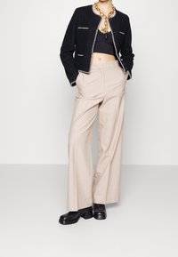 Czarna krótka kurtka z białym wykończeniem, w połączeniu z beżowymi spodniami typu wide-leg i czarnymi botkami na obcasie. Duży złoty naszyjnik jako akcent.