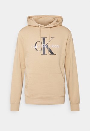 Beżowa bluza z kapturem z przednią kieszenią i sznurkami, z dużymi czarnymi literami "CK" i napisem "Calvin Klein Jeans" na piersi.
