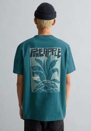 Personne aux cheveux blonds courts portant un bonnet sombre et un t-shirt bleu-vert avec un grand graphique stylisé d'ananas et du texte au dos.