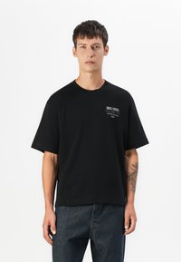 Sort bomulds t-shirt med en afslappet pasform, korte ærmer og en brystprint med hvid tekst. Båret med mørke denim jeans.