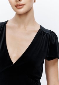 Abito in velluto nero caratterizzato da una profonda scollatura a V, maniche corte a sbuffo e vita aderente. Tessuto liscio con una leggera lucentezza.