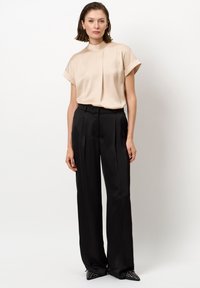 Beige Bluse mit kurzen Ärmeln und hohem Kragen; kombiniert mit schwarzen hoch taillierten Hosen; spitze schwarze Schuhe mit Nietenakzenten.