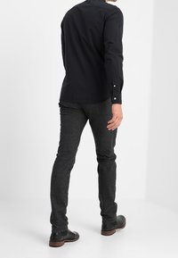 Black long-sleeve shirt with button cuffs, paired with dark grey textured trousers and black leather shoes. Rear view displayed.