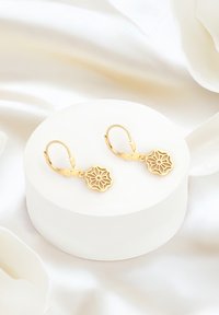 Orecchini a cerchio in oro con un design floreale intagliato complesso. Esibiti su un pedestal rotondo bianco contro uno sfondo di tessuto morbido.