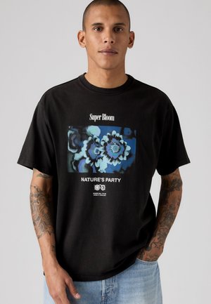 Hombre con cabello corto y brazos tatuados, de pie mirando hacia adelante, que lleva una camiseta negra con arte de flor azul y texto "Super Bloom Nature's Party."