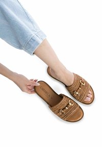 Sandalias deslizantes tejidas en tono marrón con acentos de hardware dorado. La parte superior presenta una textura trenzada y la suela es plana y de color marrón.
