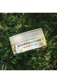 Pot transparent en forme de cercle contenant du beurre corporel, avec un couvercle texturé, arborant des motifs d'oiseaux jaunes et une étiquette avec "SUSANNE KAUFMANN" en texte noir.
