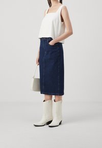 Vit cropped top med fyrkantig halsringning, mörkblå denim midi-kjol, vita cowboystövlar med svarta klackar och en liten grå handväska.