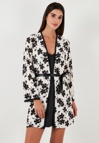 Kimono lung floral în alb și negru, cu dantelă la margine și o curea neagră. Are o croială lejeră și mâneci lungi, purtat peste o rochie neagră.