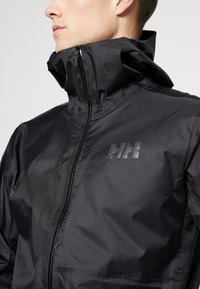 Chaqueta cortavientos negra con cuello alto, tejido texturizado, cremallera frontal y un logo sutil en el pecho. Diseño ligero y ajustado.