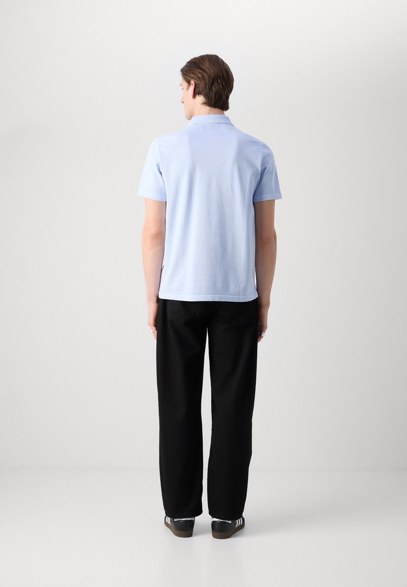 Himmelblaues Kurzarm-Poloshirt mit Kragen, kombiniert mit schwarzen, weit geschnittenen Hosen und dunklen Schuhen mit weißen Streifen, von hinten betrachtet.