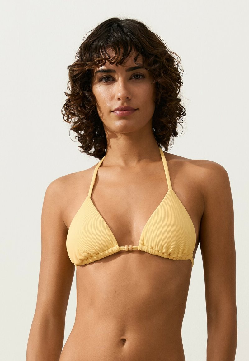 Jeune femme aux cheveux courts et bouclés portant un haut de bikini jaune à col halter, debout devant un fond clair uni.