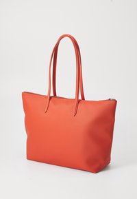 Sac fourre-tout orange en matière texturée, doté de longues poignées fines et d'une fermeture éclair en haut, avec une forme rectangulaire.