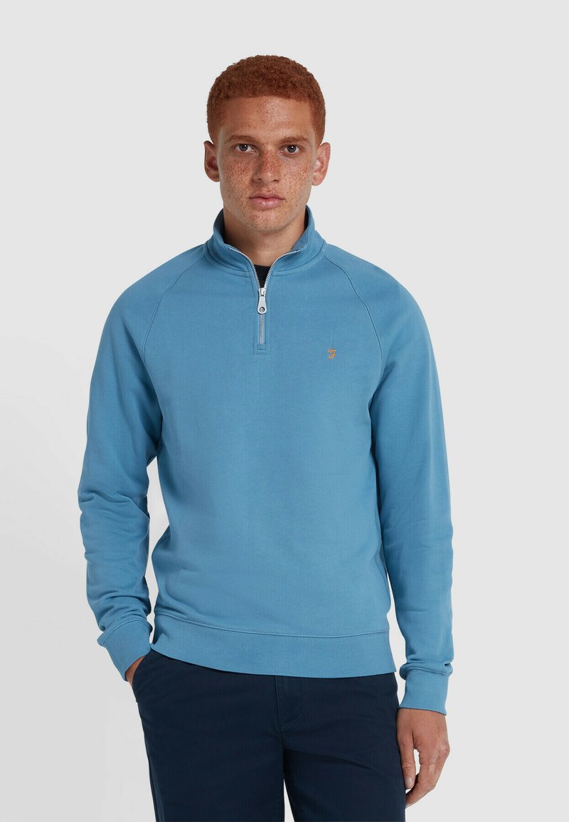 Maglione con zip blu chiaro e colletto alto, realizzato in tessuto morbido, con un piccolo logo sul petto e polsini a costine.