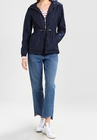 Giacca a vento blu navy con cappuccio e cordini elasticizzati, chiusura con zip e maniche lunghe, abbinata a jeans a vita alta leggeri di colore azzurro e sneakers bianche.