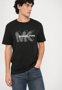 Mladý muž oblečený v čiernom tričku značky Michael Kors s veľkým logom MK, stojí s jednou rukou vo vrecku a pozerá sa na stranu.