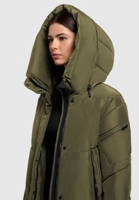 Olivovo zelená nadýchaná bunda s prehnanou kapucňou, s predným zipsom, bočnými vreckami a hladkou textúrou. Viditeľné spevnené švy a patentky.