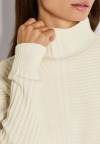 Vrouw die de kraag van een crèmekleurige, ribbelgebreide trui met lange mouwen aanpast.