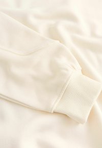 Cremefarvet, blødt strik sweatshirt med ribbet manchet, der viser en let tekstur og glat stof. Synlige sømdetaljer forbedrer designet.