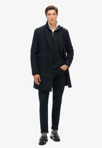 Cappotto peacoat in lana blu navy con bottoni doppi, indossato su un maglione nero, abbinato a jeans scuri e mocassini neri.