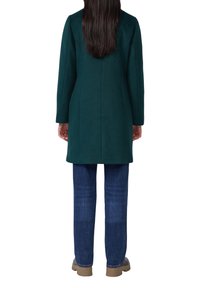 Grüner Wollmischmantel mit langen Ärmeln, strukturierten Schultern und gerader Silhouette, kombiniert mit blauen Jeans und beigen Schuhen.