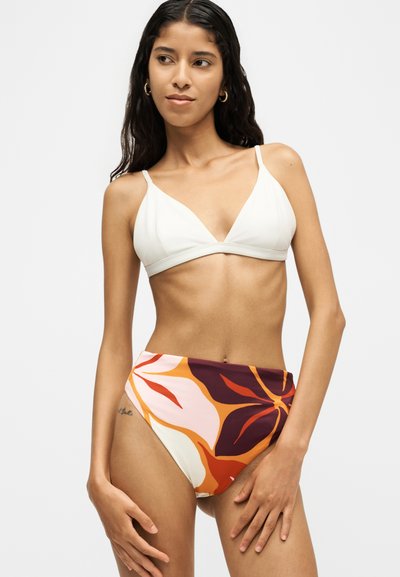 Femme aux longs cheveux foncés portant des créoles dorées, un haut de bikini blanc et un bas de bikini fleuri taille haute dans les tons orange, violet et rose.
