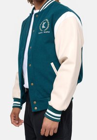 Biało-niebieska kurtka typu varsity z wełnianym tułowiem, skórzanymi rękawami, ściągaczami, złotymi zatrzaskami oraz haftowanym logo na klatce piersiowej.