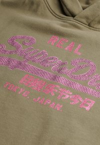 Felpa verde oliva con testo in strass rosa lucido che dice “REAL SuperDry” e “TOKYO. JAPAN” in caratteri giapponesi. Tessuto morbido.