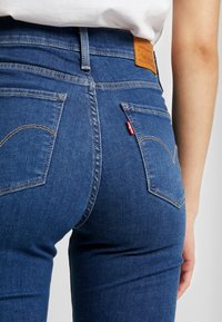 Primer plano de una persona que lleva puestos unos vaqueros de mezclilla azules con bolsillos traseros, costuras visibles y una etiqueta de cuero marrón de la marca en la cintura.