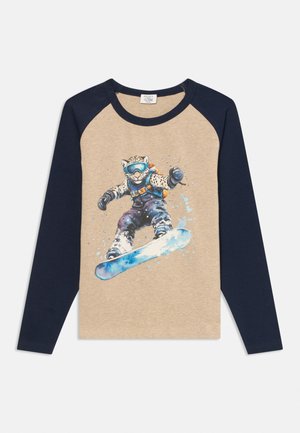 Maglietta beige e blu navy a maniche lunghe con la grafica di un leopardo che fa snowboard, indossando occhiali da sci e abbigliamento invernale, a metà salto su uno snowboard blu.