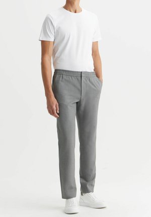 Mand iført en enkel hvid t-shirt med korte ærmer, grå bukser med lige ben og elastisk linning samt hvide sneakers, står med hænderne i lommerne.
