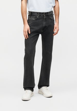 Férfi kopott fekete egyenes szárú farmernadrágot, fehér tornacipőt és világoskék, betűrt inget visel, fehér háttér előtt áll.