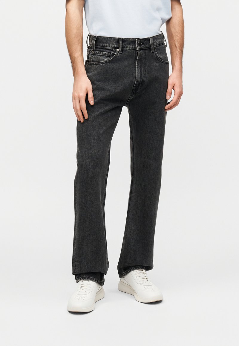 Άνδρας που φοράει ξεθωριασμένο μαύρο τζιν με ίσια μπάρα, λευκά αθλητικά παπούτσια και ανοιχτό μπλε πουκάμισο μπουκαρισμένο μέσα στο παντελόνι, στέκεται μπροστά σε λευκό φόντο.