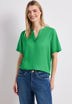 Femme souriante aux longs cheveux blonds portant un chemisier vert à manches courtes et un jean bleu, debout devant un fond uni.