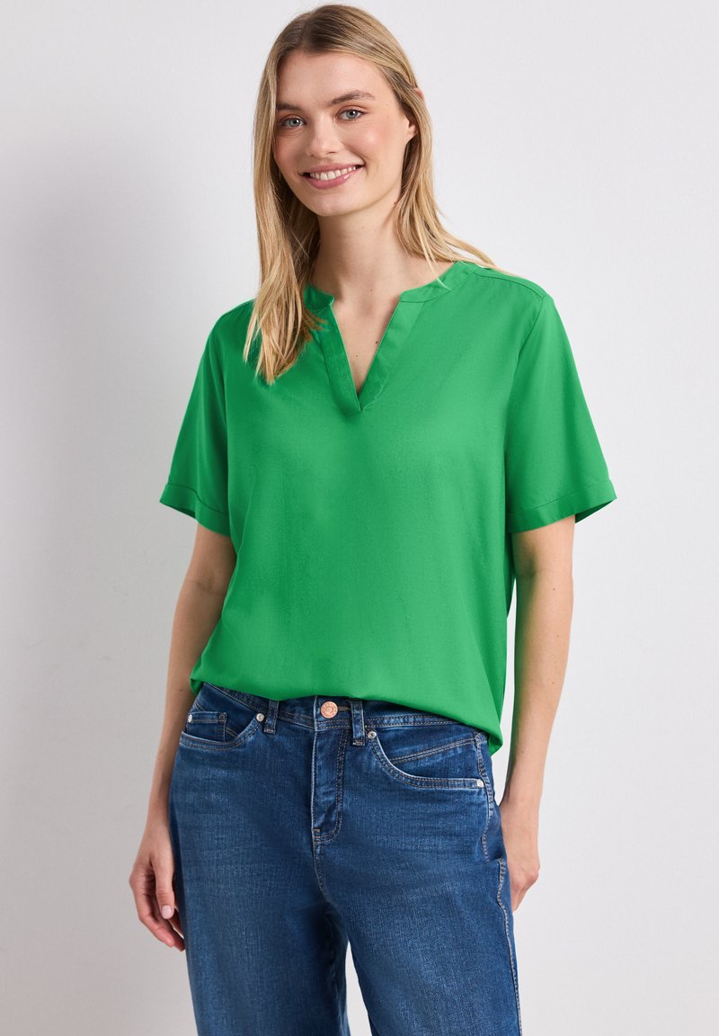 Femme souriante aux longs cheveux blonds portant un chemisier vert à manches courtes et un jean bleu, debout devant un fond uni.