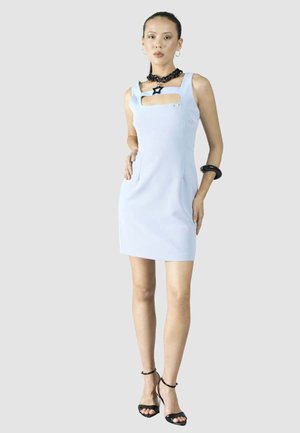 Donna che indossa un vestito azzurro chiaro senza maniche con apertura sul petto, collana con stella nera, braccialetto nero e sandali neri con tacco alto, in piedi.