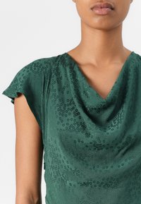 Green satin top with a draped neckline, short flutter sleeves, and a textured pattern featuring small circles throughout.