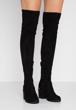 Bottes over-the-knee en daim noir avec talons épais portées sur des jambes nues sur un fond gris clair uni.