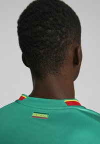 Turkis sportstrøje med en rød og gul stribet krave, der har et "SENEGAL" mærke på bagsiden. Glat stoftekstur.