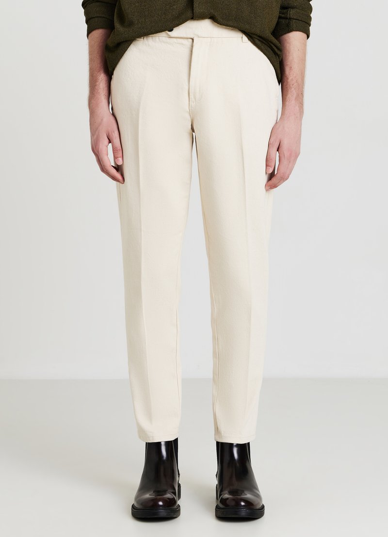 Cream-colored trousers with a textured finish, tailored fit, and side pockets, paired with glossy black ankle boots.