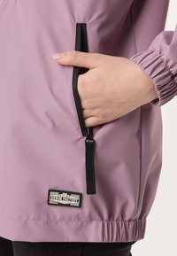 Hand partially inside front zipper pocket of mauve jacket with elastic cuff and Denim Factory label near hem.