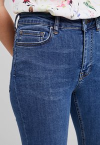 Primo piano di una persona che indossa dei jeans in denim blu a vita alta con cuciture arancioni e una blusa floreale bianca infilata dentro.