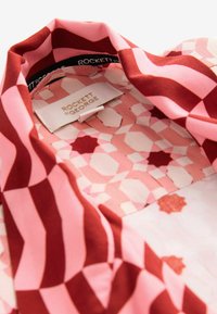 Raštuotas audinys, turintis pakaitomis rožinių ir raudonų atspalvių, su geometriniais piešiniais. Ant jo yra etiketė su užrašu "ROCKETT & GEORGE."