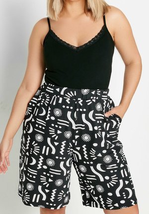 Frau trägt ein schwarzes Camisole mit Spitzenbesatz und schwarze Shorts mit weißen abstrakten Sonnen- und Mondmustern.