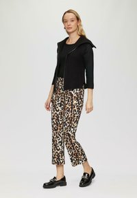 Czarna dzianinowa kamizelka z kapturem na czarnym topie, zestawiona z spodniami w leopardzi wzór w odcieniach beżu i brązu, dopełniona czarnymi loafersami.