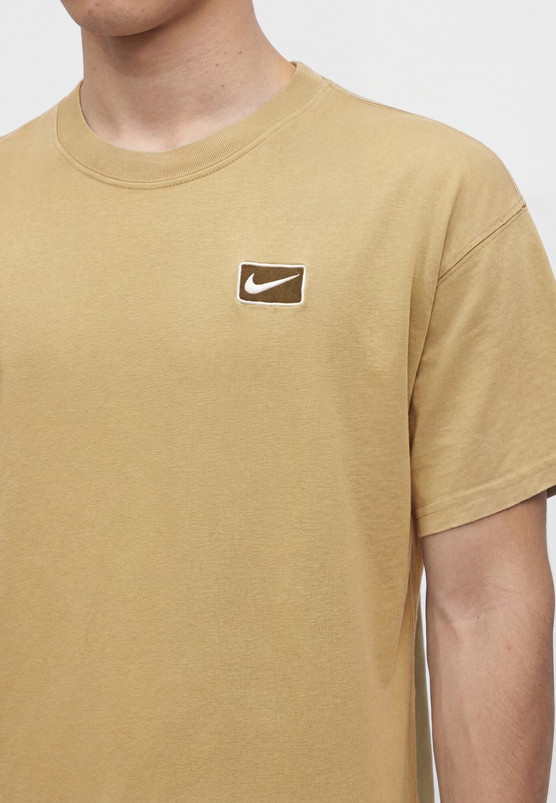 Mann trägt beige kurzärmeliges T-Shirt mit kleinem braun-weißem Nike-Logo auf der linken Brust.