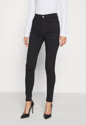 Jeans skinny de cintura alta negros con diseño de cinco bolsillos y cierre de botón. Combinados con tacones negros de punta afilada y una blusa blanca de manga larga.