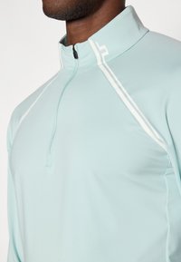 Gaiši zils ar rāvējslēdzēju augšpusē, garām piedurknēm tops ar augstu apkakli, ar baltām akcenta krāsām un gludu, elastīgu audumu.